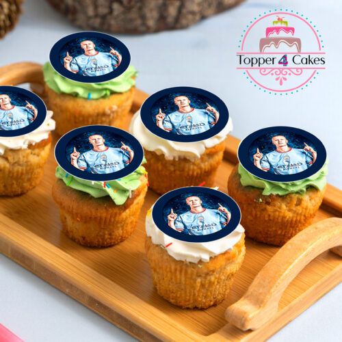 A wooden tray displays six cupcakes, each embellished with edible images of a man wearing a light blue sports shirt and topped with green and white frosting. In the background, a logo for Topper 4 Cakes is visible next to a cake illustration, showcasing their personalised Edible Liverpool Cupcake Topper Personalised product.