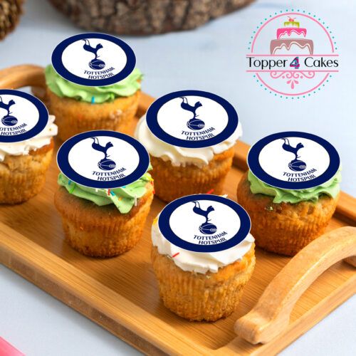 A wooden tray displays six cupcakes adorned with white and green frosting, each featuring an edible Liverpool cupcake topper. In the background, a pink circular sign with the Topper 4 Cakes logo subtly promotes their personalised Liverpool cake decorations, perfect for delighting every fan.