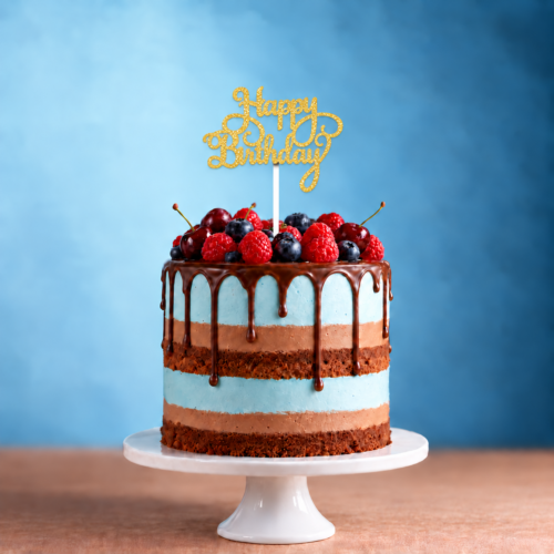 A layered chocolate cake with blue icing, topped with fresh berries and the Happy Birthday Glitter Cake Topper, rests on a white cake stand against a blue background.