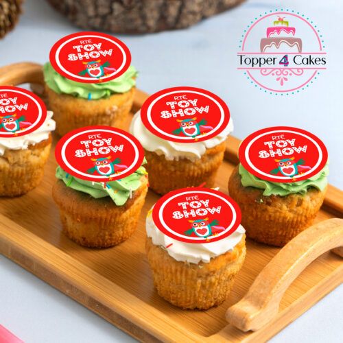 A wooden tray holds six Christmas cupcakes, each with green or white frosting and a red topper featuring a cartoon owl and RTÉ Toy Show. The Topper 4 Cakes logo is prominent in the top right corner, perfect for The Late Late Toy Show Cupcake Toppers collection.