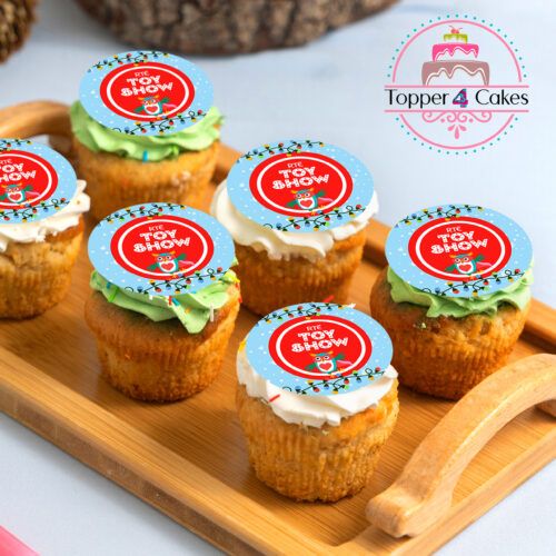 A wooden tray holds six Christmas cupcakes with cream frosting, each topped with a circular design featuring festive themes and Toy Show. In the background, the logo for The Late Late Toy Show Cupcake Toppers adds a charming touch.