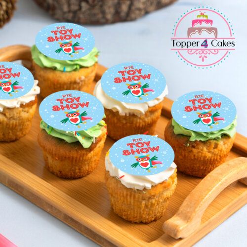 A wooden tray features six Christmas cupcakes, each decorated with vibrant blue edible toppers featuring cartoon characters and the text RTÉ Toy Show. The diverse frosting colors of these festive treats contrast beautifully against a light-toned surface.