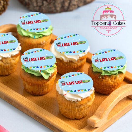 Six Christmas cupcakes with colorful frosting and edible RTE The Late Late decorations, featuring The Late Late Toy Show Cupcake Toppers, are displayed on a wooden tray. A Topper 4 Cakes logo can be seen in the background.